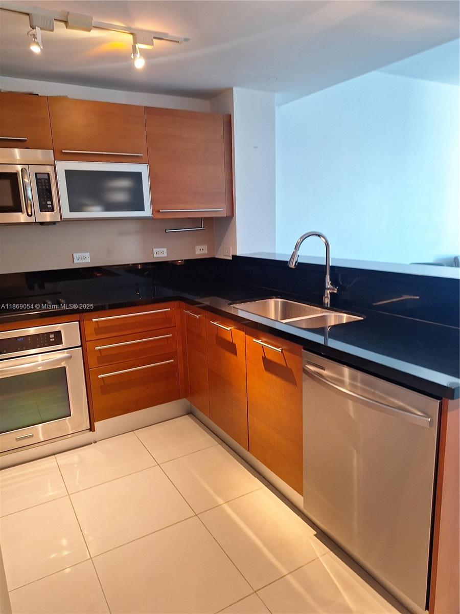 A kitchen features wood cabinets, black countertops, stainless steel appliances, and light-colored tile flooring — 253 NE 2nd St # 2606, Miami FL 33132, MIAMI, FL