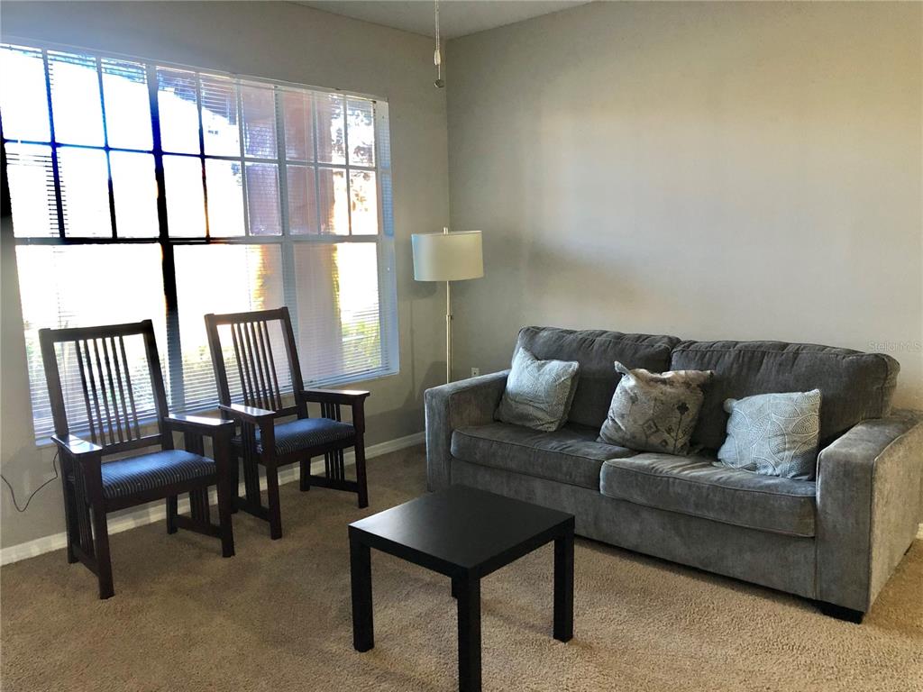 A living room features a gray sofa, two wooden chairs, a coffee table, and windows — 5542 METROWEST BOULEVARD UNIT 104, ORLANDO, FL