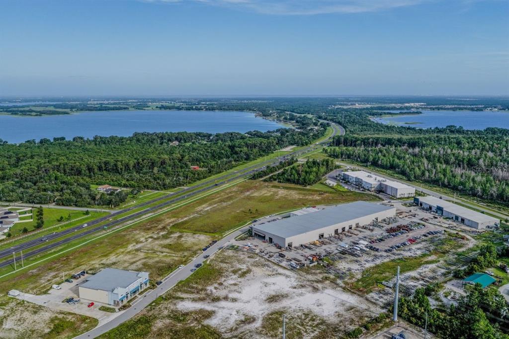 An aerial view shows commercial warehouse buildings, a paved road, and surrounding forest and water — 0 E IRLO BRONSON MEMORIAL HIGHWAY, ST CLOUD, FL