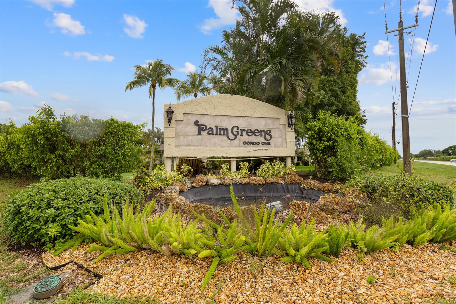 A stone sign for Palm Greens Condo One sits behind a landscaped garden and pond — 13623 Via Aurora # C, Delray Beach FL 33484, DELRAY BEACH, FL