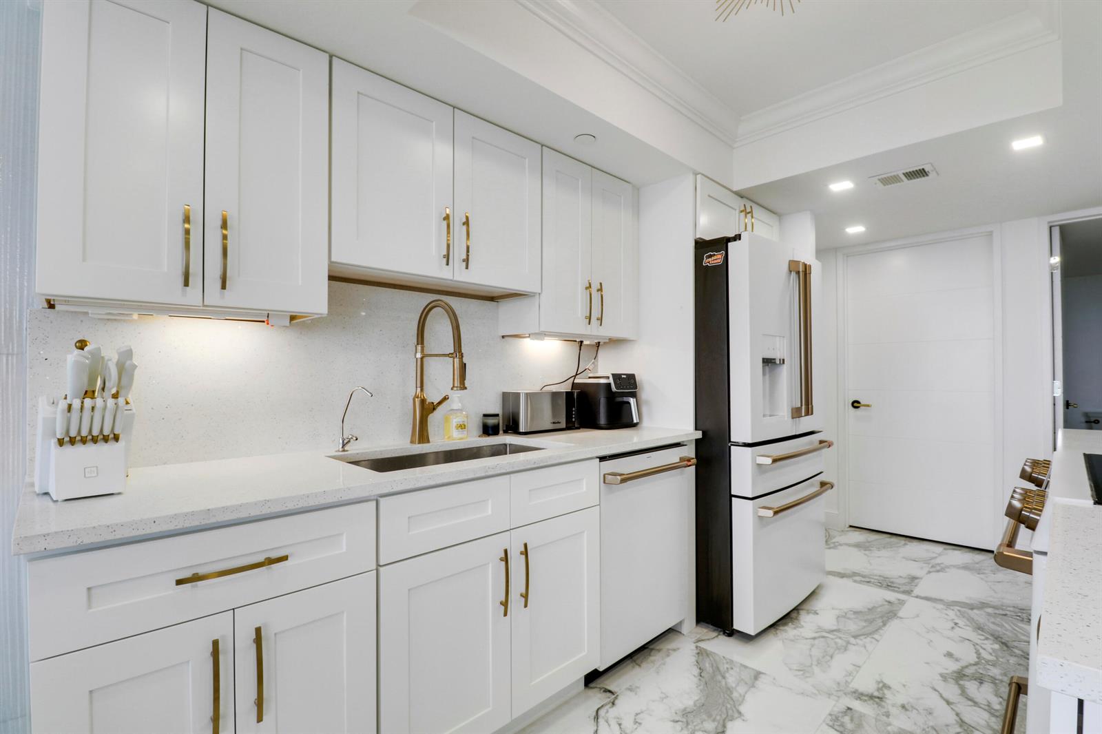 A modern kitchen features white cabinets, gold hardware, a stainless sink, and marble-patterned tile flooring — 5280 N Ocean Drive # 2f, Riviera Beach FL 33404, RIVIERA BEACH, FL