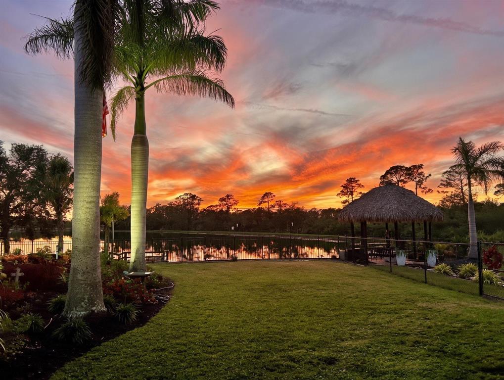 A grassy backyard features a palm tree, a tiki hut, and a lake at sunset — 9150 DEER COURT, VENICE, FL