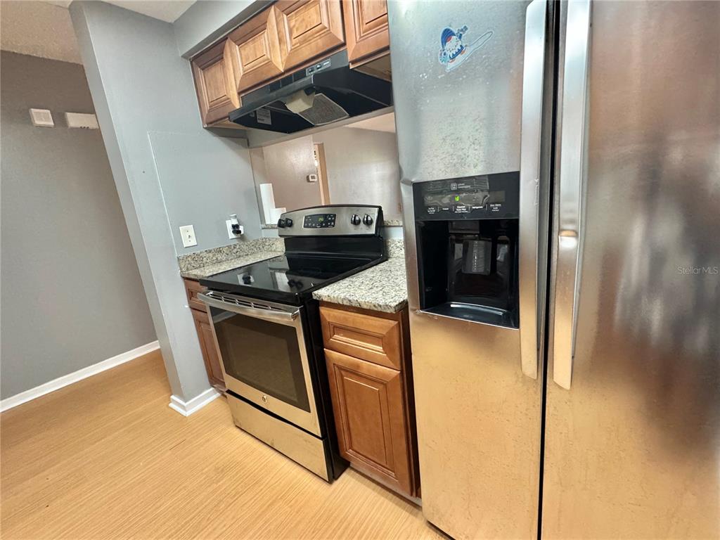 A kitchen features stainless steel appliances, wood cabinets, and granite countertops over light wood flooring — 4852 MARKS TERRACE UNIT 52, ORLANDO, FL
