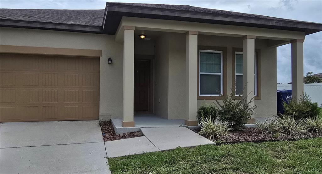 The exterior of a single-story house features a tan stucco facade, a garage, and columns — 15660 MERLIN AVENUE, MASCOTTE, FL