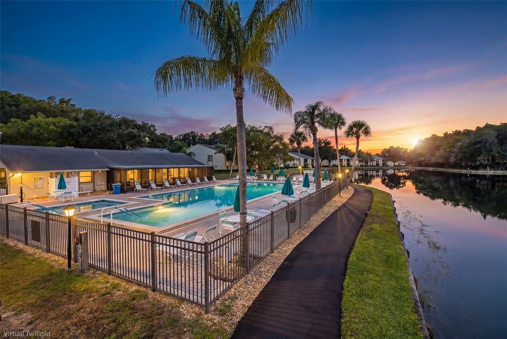 An outdoor swimming pool and patio area sit beside a lake at sunset — 3166 LAKE PINE WAY S UNIT A2, TARPON SPRINGS, FL