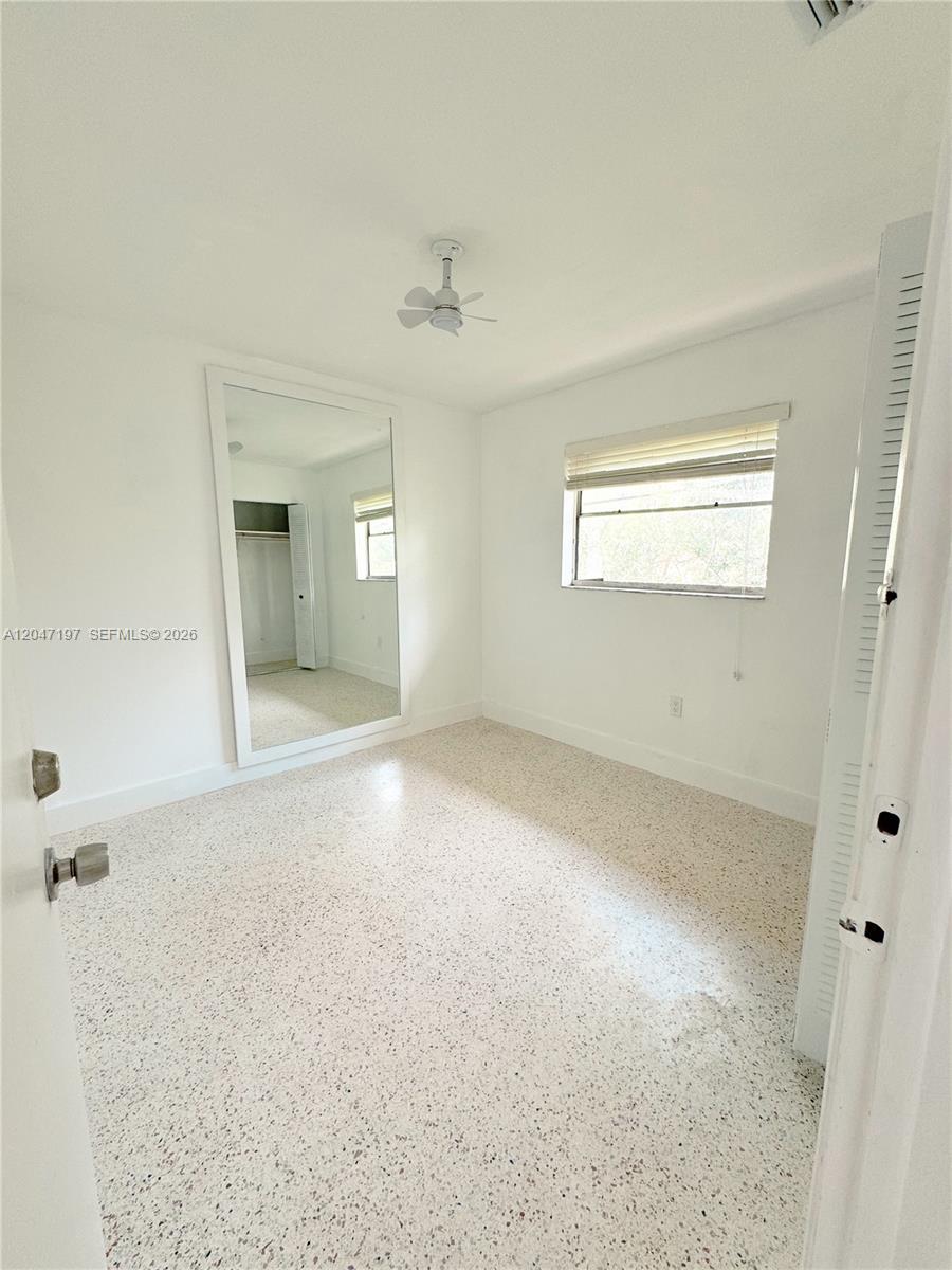 A bedroom features white walls, a ceiling fan, a large mirror, and speckled flooring — MIAMI, FL