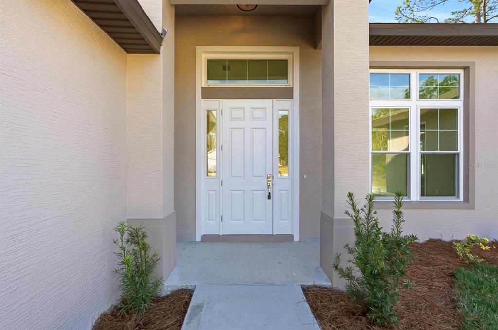 A white front door with sidelights and a transom window sits under a covered entryway — 16 PIERCE LANE, PALM COAST, FL