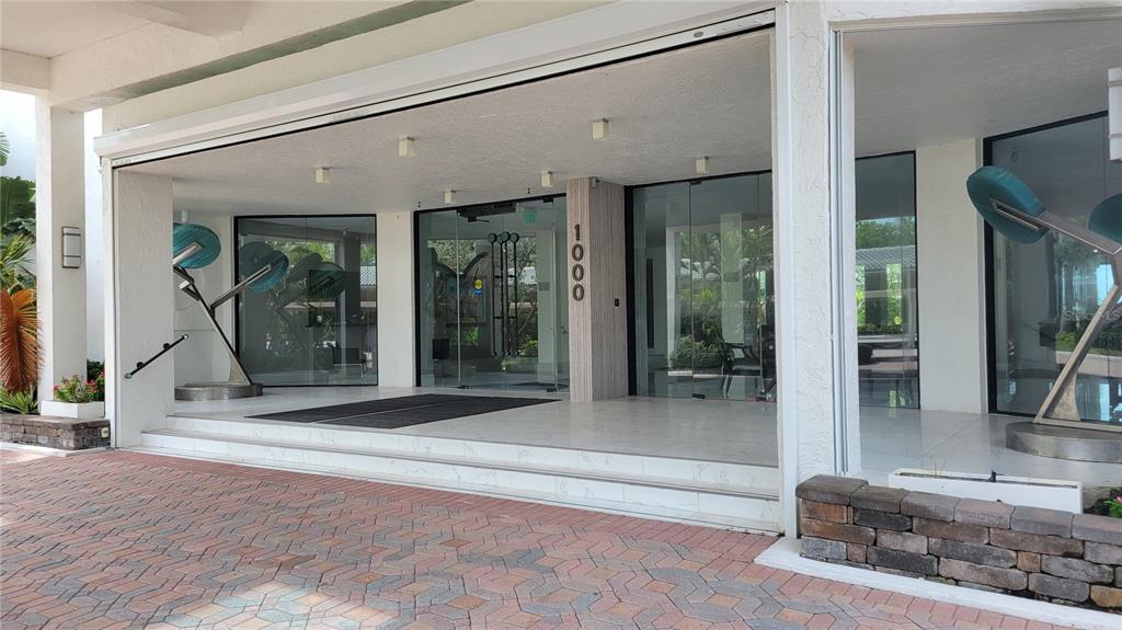 The exterior entrance of a building features glass doors, a brick driveway, and metal sculptures — 1000 LONGBOAT CLUB ROAD UNIT 406, LONGBOAT KEY, FL