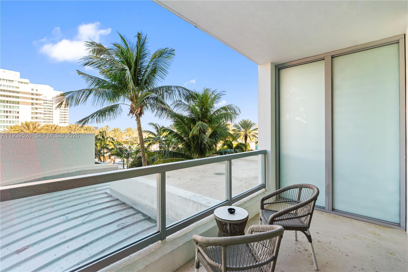 A balcony features two woven chairs, a small table, and a glass railing overlooking palms — 4391 Collins Ave # 303, Miami Beach FL 33140, MIAMI BEACH, FL