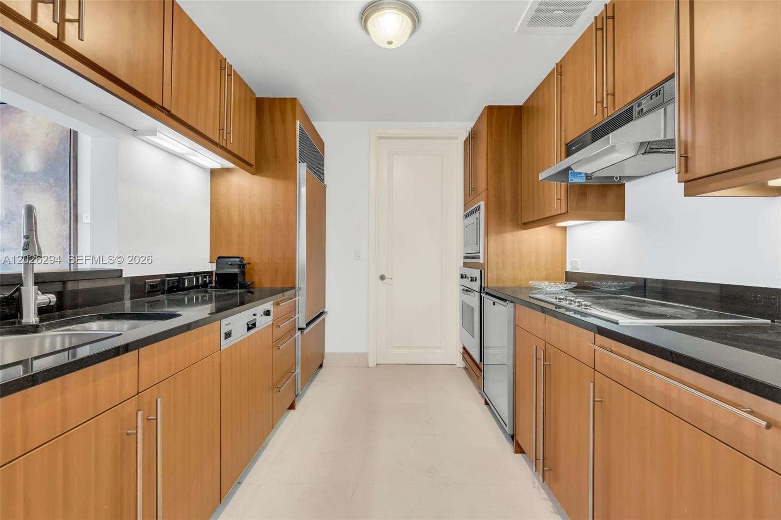 A galley kitchen features wood cabinetry, black countertops, stainless steel appliances, and light tile flooring — 1425 Brickell Ave # 51A, Miami FL 33131, MIAMI, FL