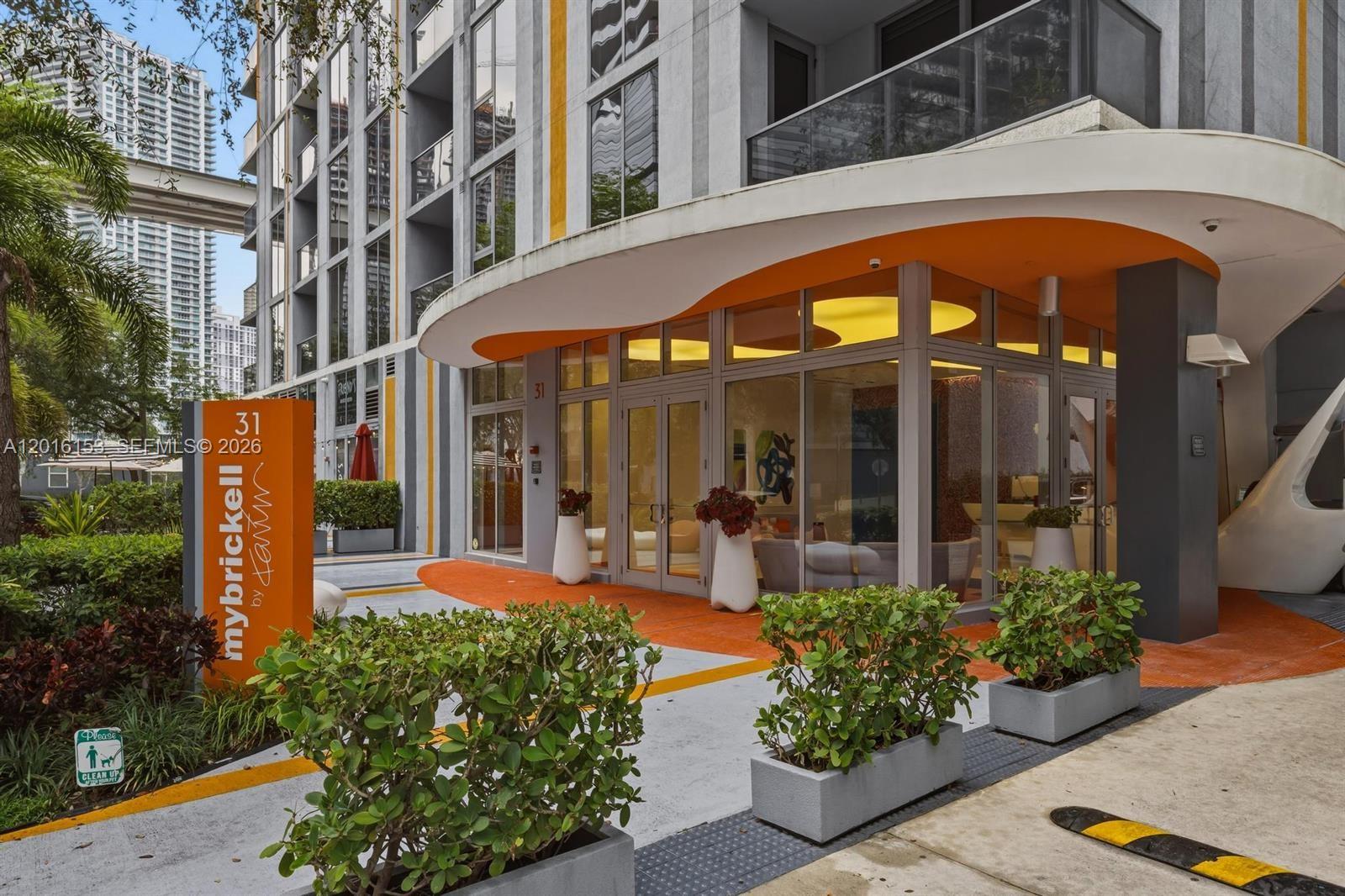 The exterior entrance of a modern apartment building features orange accents, glass doors, and planters — 31 SE 6th St # 1904, Miami FL 33131, MIAMI, FL