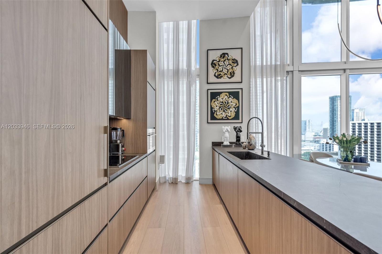 A modern galley kitchen features light wood cabinetry, dark countertops, and floor-to-ceiling windows — 475 Brickell Ave # 2215, Miami FL 33131, MIAMI, FL