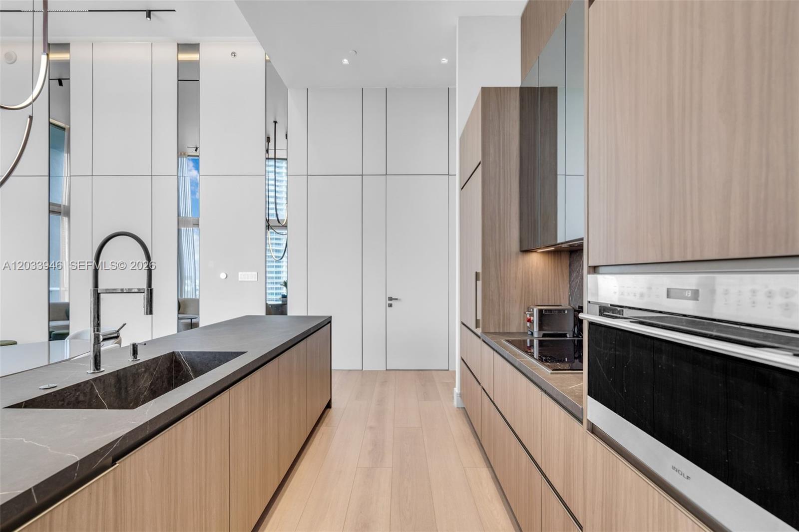 A modern kitchen features wood-toned cabinetry, a dark stone island, and high-ceiling white walls — 475 Brickell Ave # 2215, Miami FL 33131, MIAMI, FL