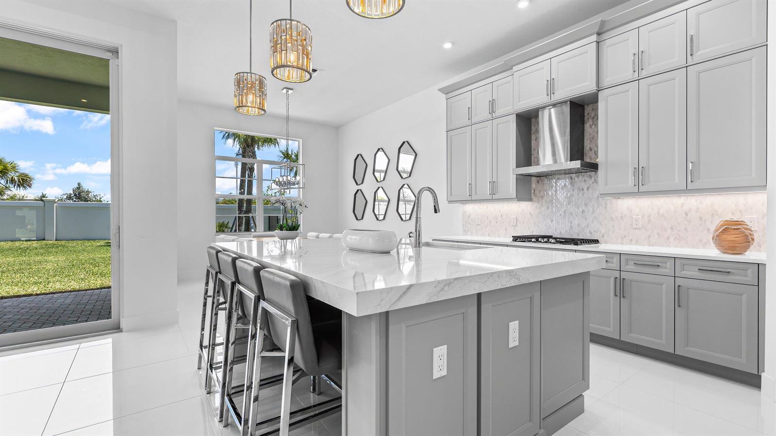 A modern kitchen features a large marble island, gray cabinetry, and a sliding glass door — 9508 St. Germain, Palm Beach Gardens FL 33412, PALM BEACH GARDENS, FL