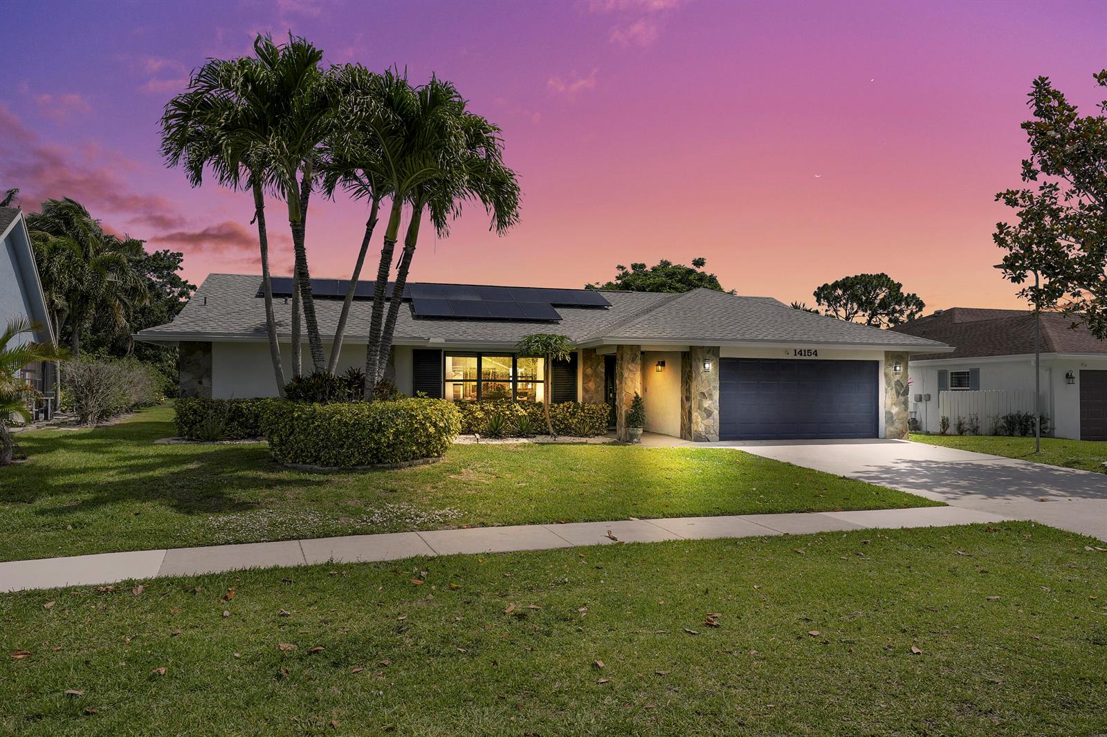 A single-story house with a dark garage door, solar panels, and palm trees at sunset — 14154 Aster Avenue, Wellington FL 33414, WELLINGTON, FL