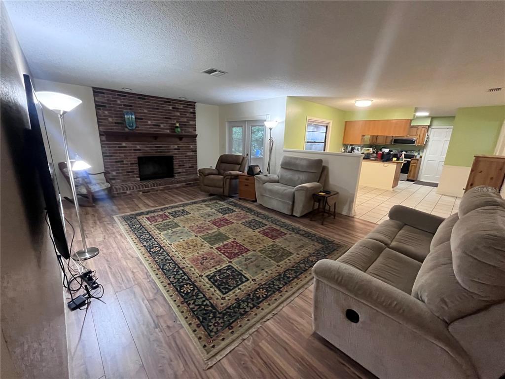 A living room with a brick fireplace, wood-look flooring, and an adjacent open kitchen — 2117 YORKSHIRE DRIVE, DELAND, FL
