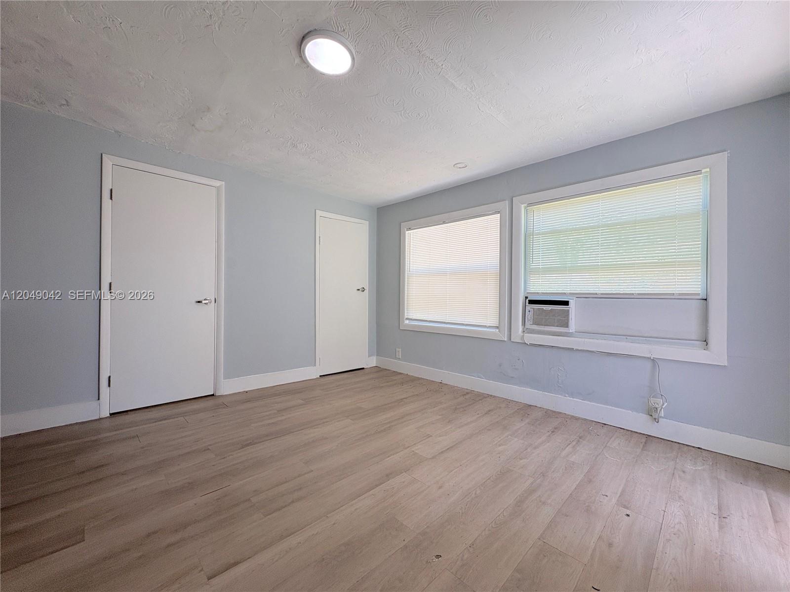 An empty room features light blue walls, light wood-look flooring, two doors, and two windows — 1825 Mckinley St # 2, Hollywood FL 33020, HOLLYWOOD, FL