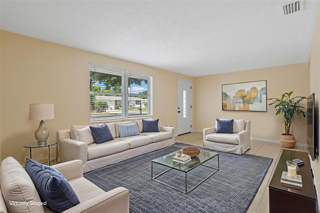 A virtually staged living room features a beige sofa, armchair, glass coffee table, and area rug — 2187 DIXIE GARDEN LOOP, HOLIDAY, FL