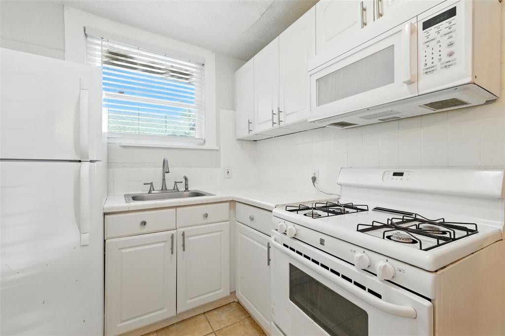 A small kitchen features white cabinets, white appliances, a tiled backsplash, and a window — 1040 19TH AVENUE N, ST PETERSBURG, FL