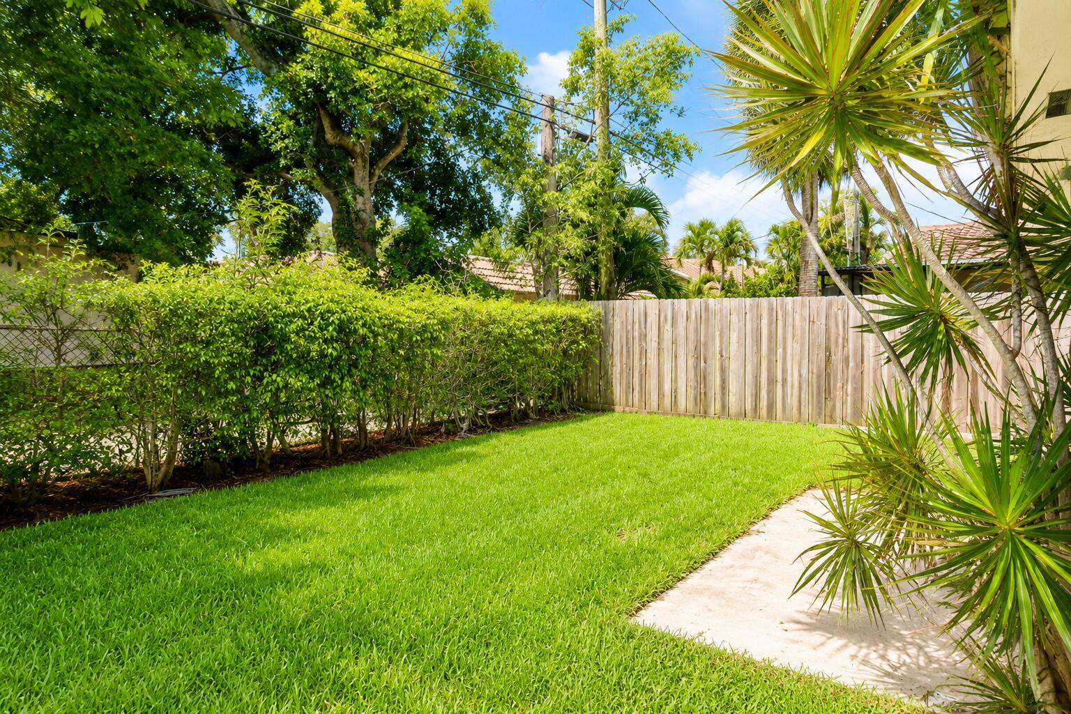 A grassy backyard features a wooden privacy fence, a hedge, and a small concrete patio — 92 Lariat Circle, Boca Raton FL 33487, BOCA RATON, FL