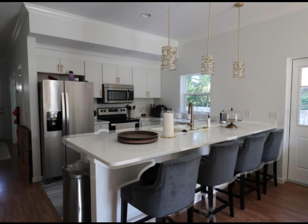 A kitchen features white cabinets, stainless steel appliances, a quartz island, and four gray barstools — 1217 ECSTASY STREET E, LEHIGH ACRES, FL