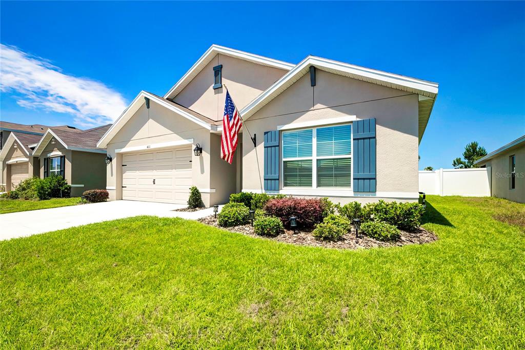 A single-story suburban house with a tan exterior, blue shutters, and an American flag — 4111 AUTUMN AMBER DRIVE, SPRING HILL, FL