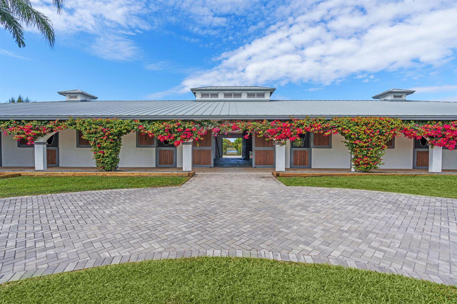 A single-story horse stable features a paved driveway, white walls, and vibrant bougainvillea climbing vines — 13155 Southfields Road, Wellington FL 33414, WELLINGTON, FL