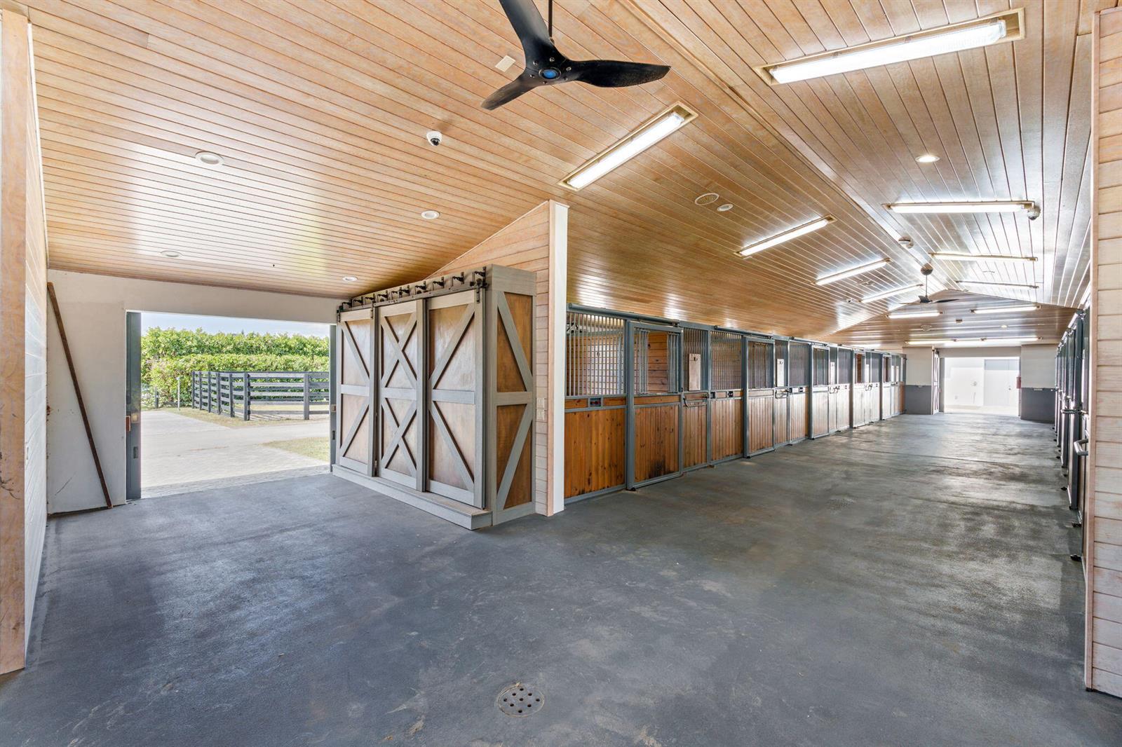 A horse stable interior features wood-paneled ceilings, concrete floors, and a row of stalls — 13155 Southfields Road, Wellington FL 33414, WELLINGTON, FL