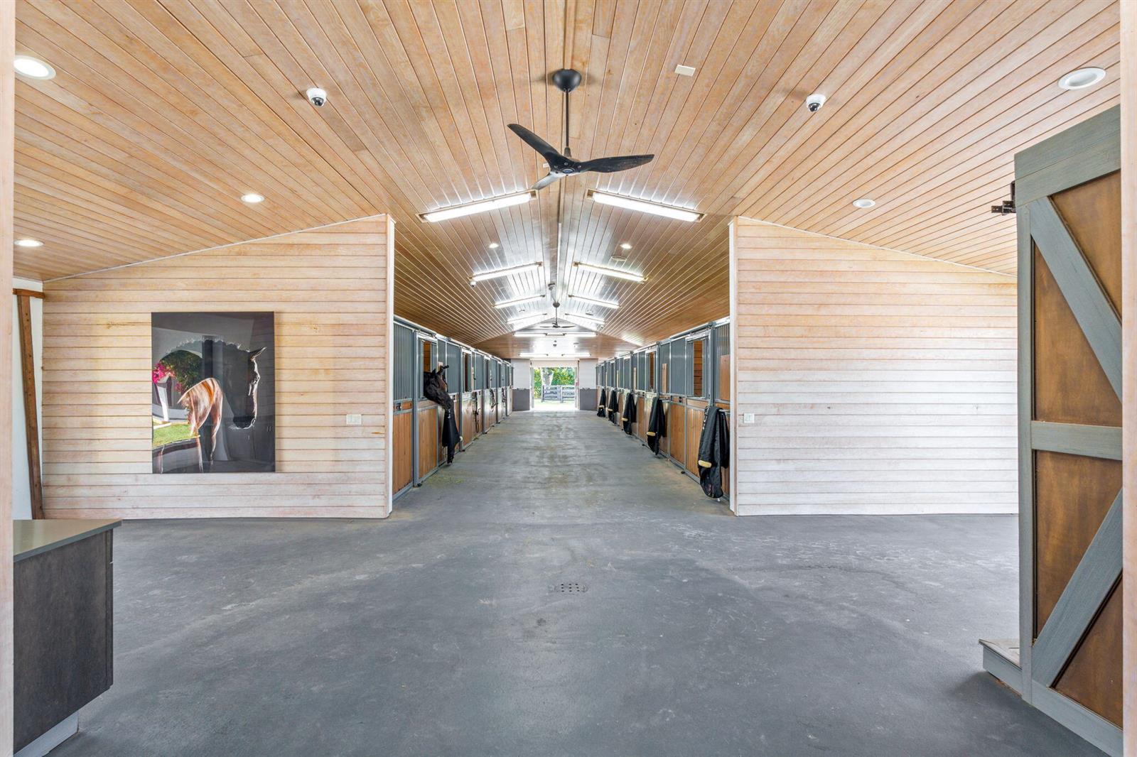 A horse stable aisle features wood-paneled walls and ceilings, concrete flooring, and multiple stalls — 13155 Southfields Road, Wellington FL 33414, WELLINGTON, FL