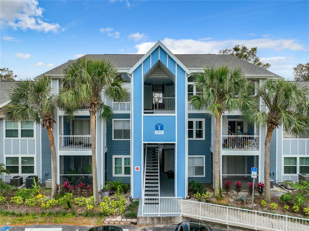 A three-story blue apartment building features a central staircase and palm trees in front — 2612 GRASSY POINT DRIVE UNIT 102, LAKE MARY, FL