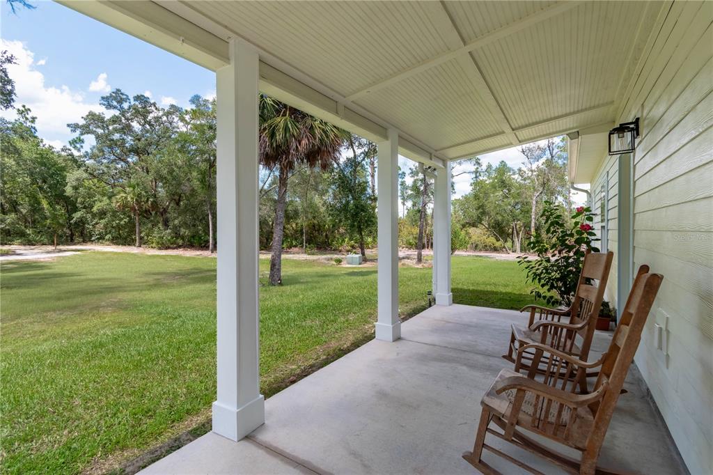A covered concrete porch features white support columns, two wooden rocking chairs, and a grassy yard — 14042 NE 110TH STREET, FORT MC COY, FL