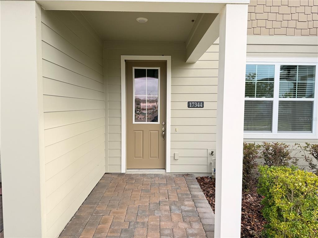 A tan front door with a window sits under a covered porch with paver flooring — 17344 SWEET MORO DRIVE, WINTER GARDEN, FL