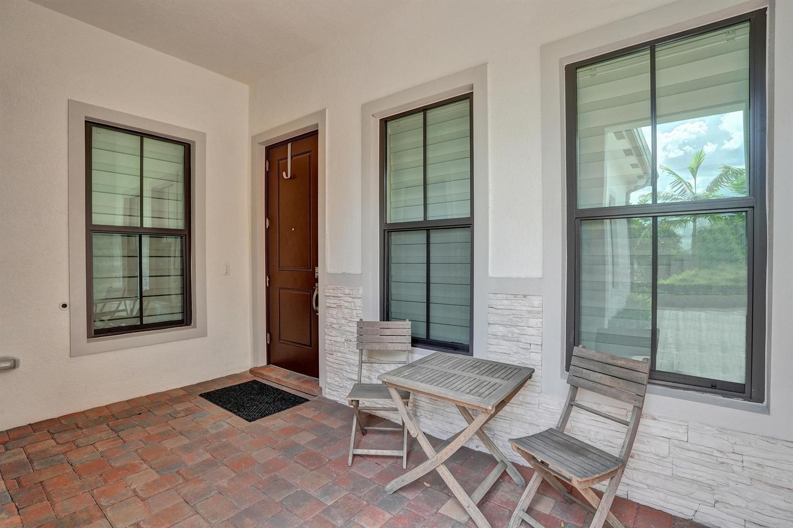 A covered patio features brick flooring, a dark wood door, and two wooden chairs — 8336 W Blue Spring Drive, Parkland FL 33067, PARKLAND, FL
