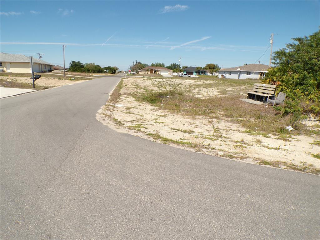 A paved street curves past a vacant sandy lot with a wooden bench — 821 NE 15TH LANE, CAPE CORAL, FL