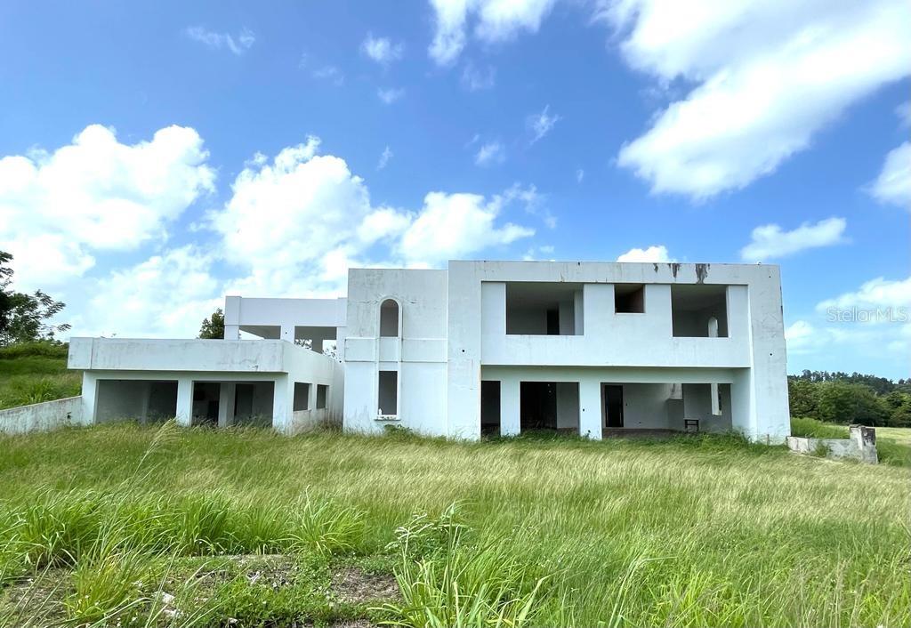 An unfinished white concrete house stands in a field of tall grass under a blue sky — PR 311 KM 2.8 BO. GUANAJIBO SECTOR LAS DELICIAS, CABO ROJO, PR