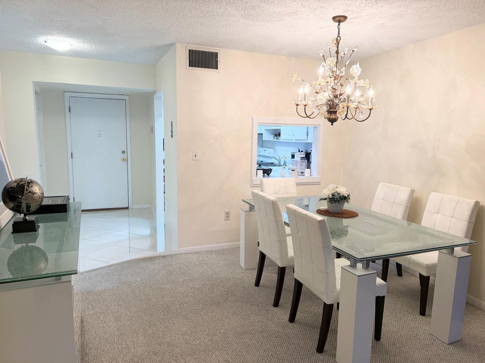 A dining room features a glass-topped table, white chairs, a crystal chandelier, and carpeted flooring — 3605 S Ocean Boulevard # A433, South Palm Beach FL 33480, SOUTH PALM BEACH, FL
