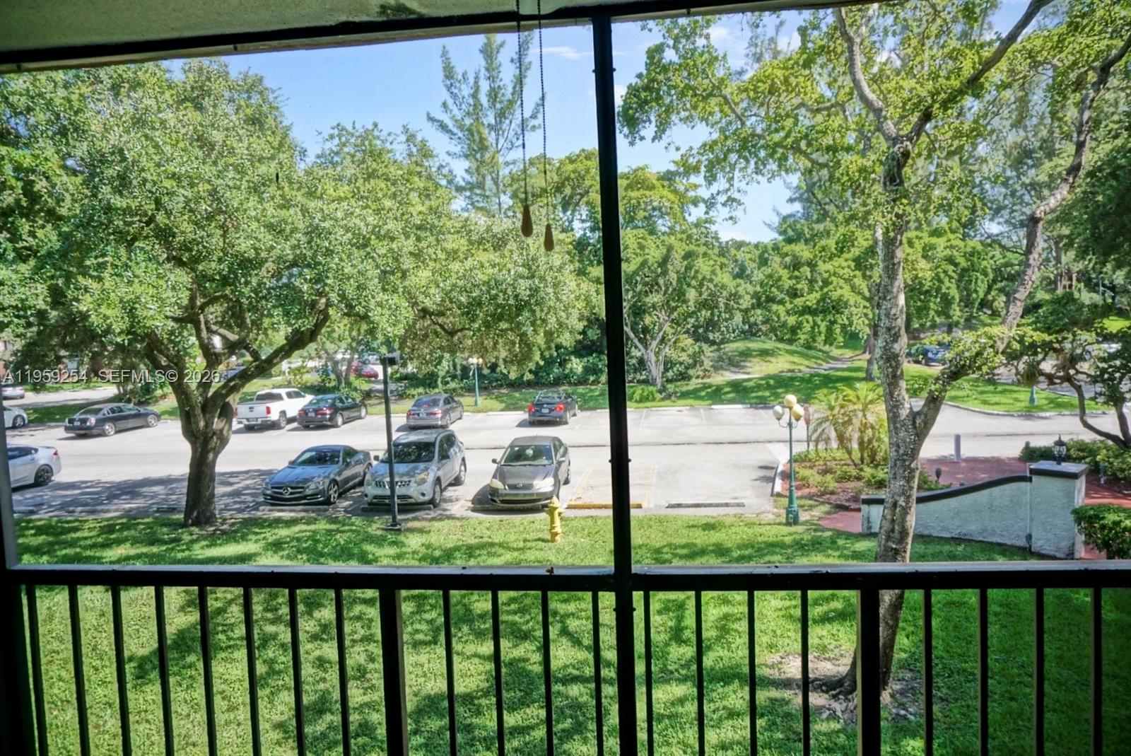 A view from a screened balcony overlooking a parking lot, green lawn, and mature trees — 3740 Inverrary Dr # 2N, Lauderhill FL 33319, LAUDERHILL, FL
