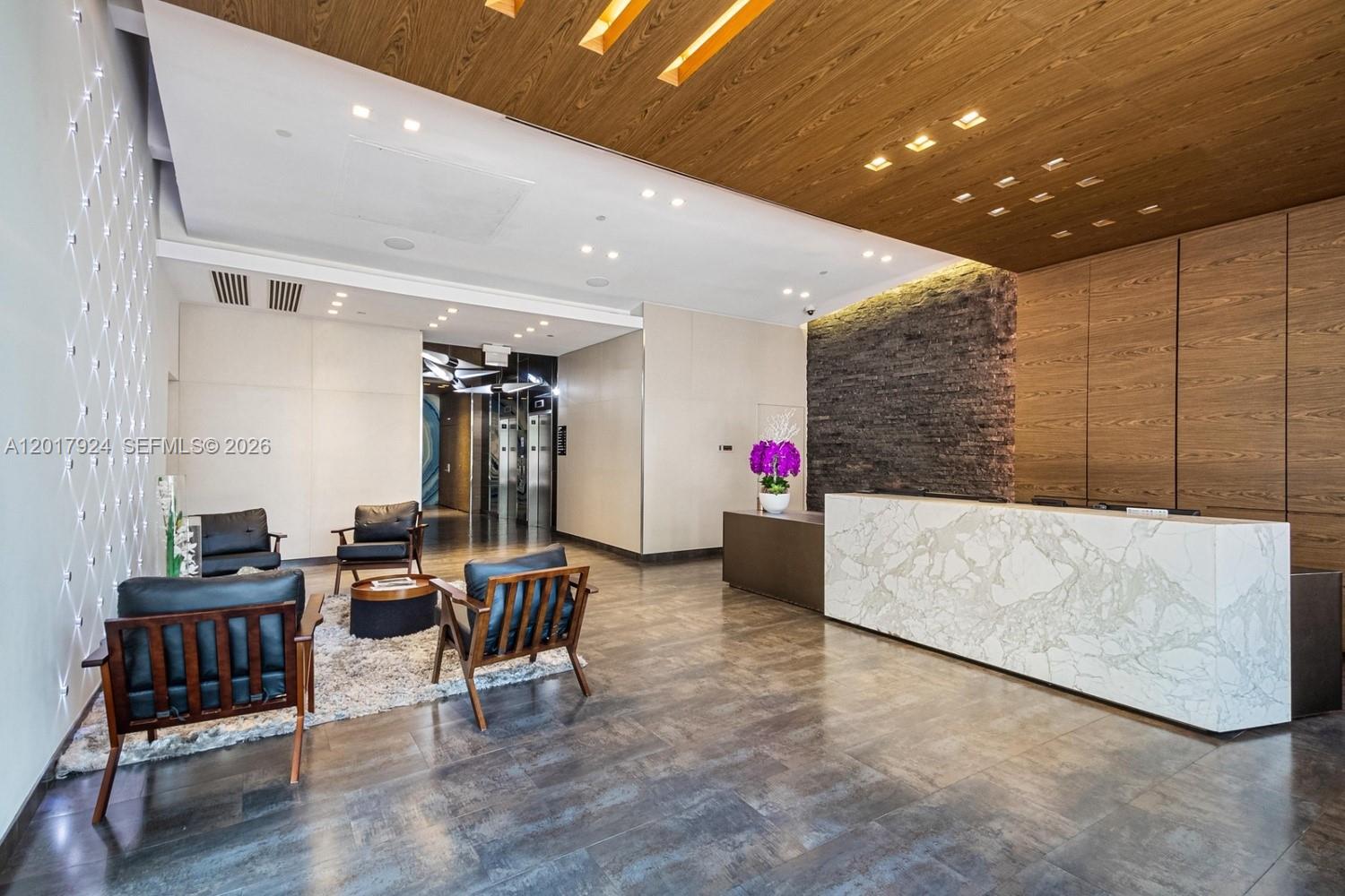 A modern lobby features a marble reception desk, wood-paneled ceiling, and a seating area — 3250 NE 1st Ave # 916, Miami FL 33137, MIAMI, FL