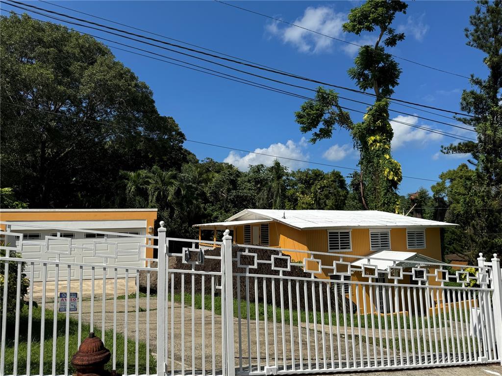 A yellow house and a detached garage are viewed from behind a white metal fence — 17 G23 COLINAS DEL YUNQUE, RIO GRANDE, PR