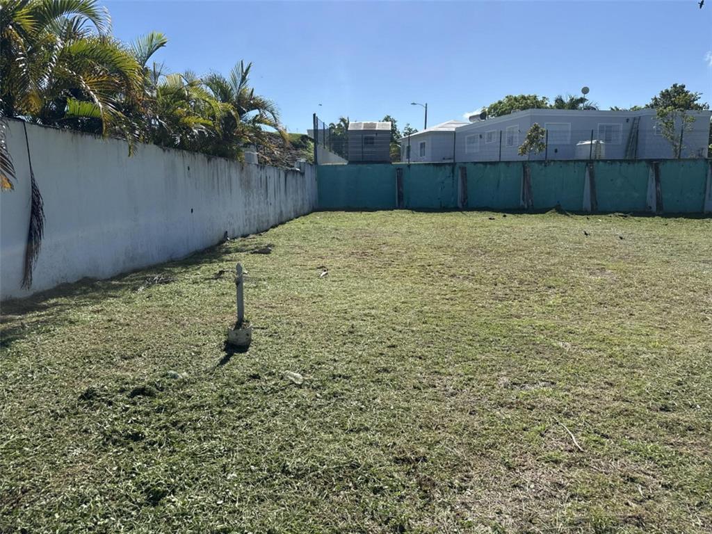 A vacant, grassy lot is enclosed by a white wall on the left and teal fencing — 7 VIENA UNIT 21, TOA ALTA, PR