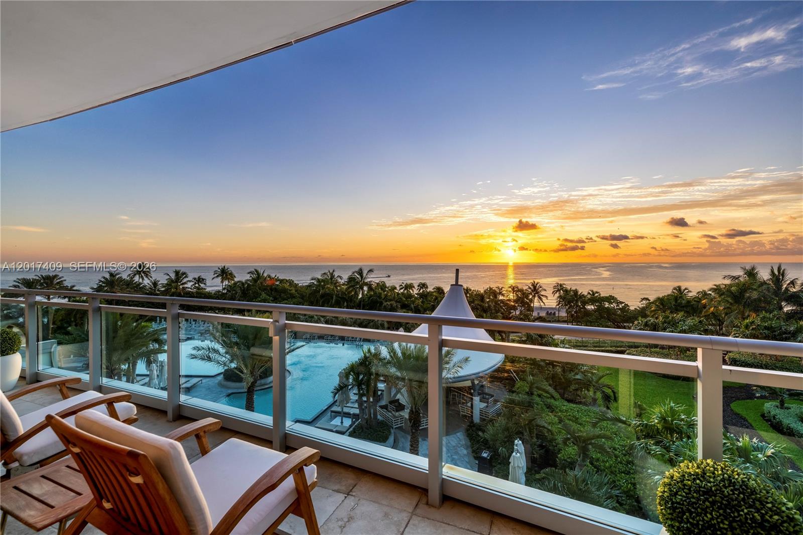 A balcony with wooden chairs overlooks a swimming pool, palm trees, and the ocean at sunset — 10295 Collins Ave # 301, Bal Harbour FL 33154, BAL HARBOUR, FL