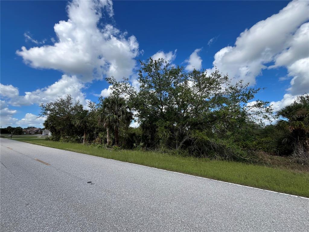 A paved road runs alongside a vacant, tree-covered lot under a blue sky with clouds — 17174 EDGEWATER DRIVE, PORT CHARLOTTE, FL