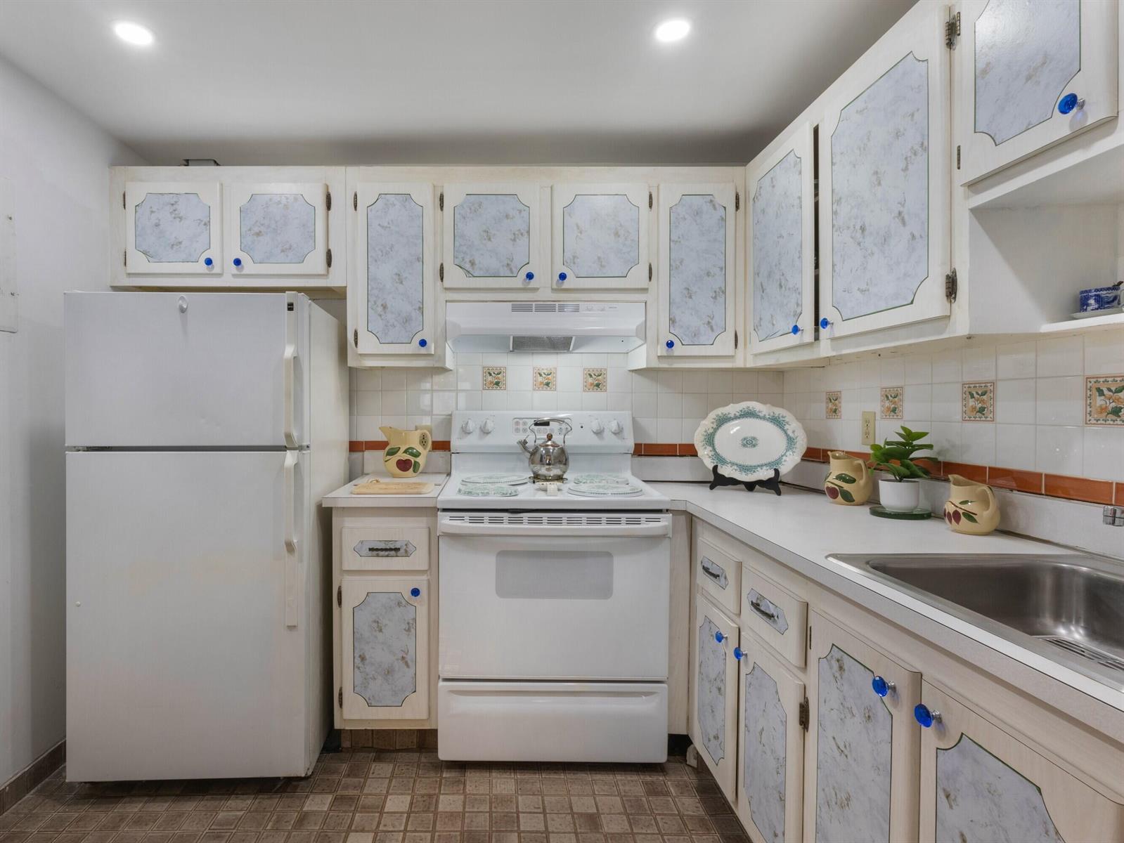 A kitchen features white appliances, patterned cabinet panels, tiled walls, and a stainless steel sink — 499 Saxony K, Delray Beach FL 33446, DELRAY BEACH, FL