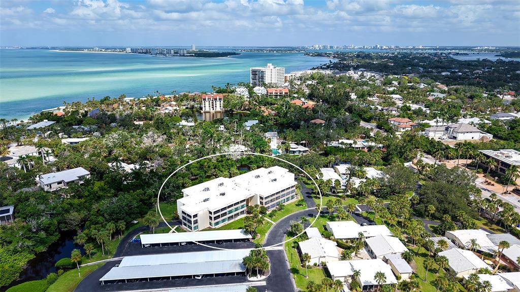 An aerial view shows a multi-story white apartment building surrounded by lush trees and parking — 225 HOURGLASS WAY UNIT 103, SARASOTA, FL