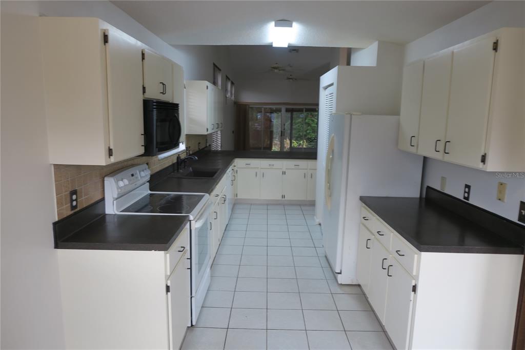 A galley kitchen features white cabinets, dark countertops, white appliances, and light-colored tile flooring — 32512 OAK PARK DRIVE, LEESBURG, FL