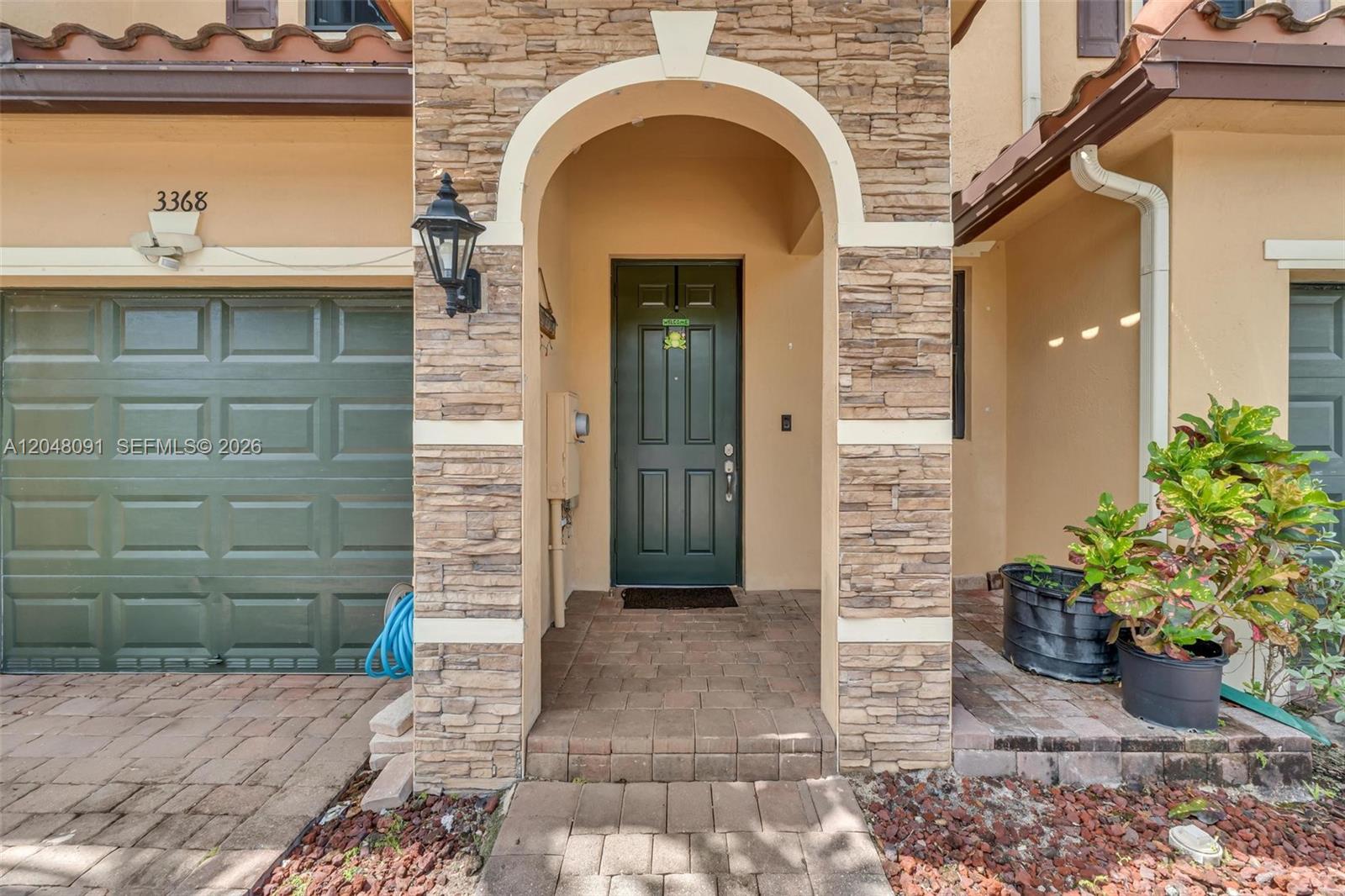 The exterior features a stone-faced arched entryway, a green garage door, and a paved walkway — 3368 SE 1st St, Homestead FL 33033, HOMESTEAD, FL