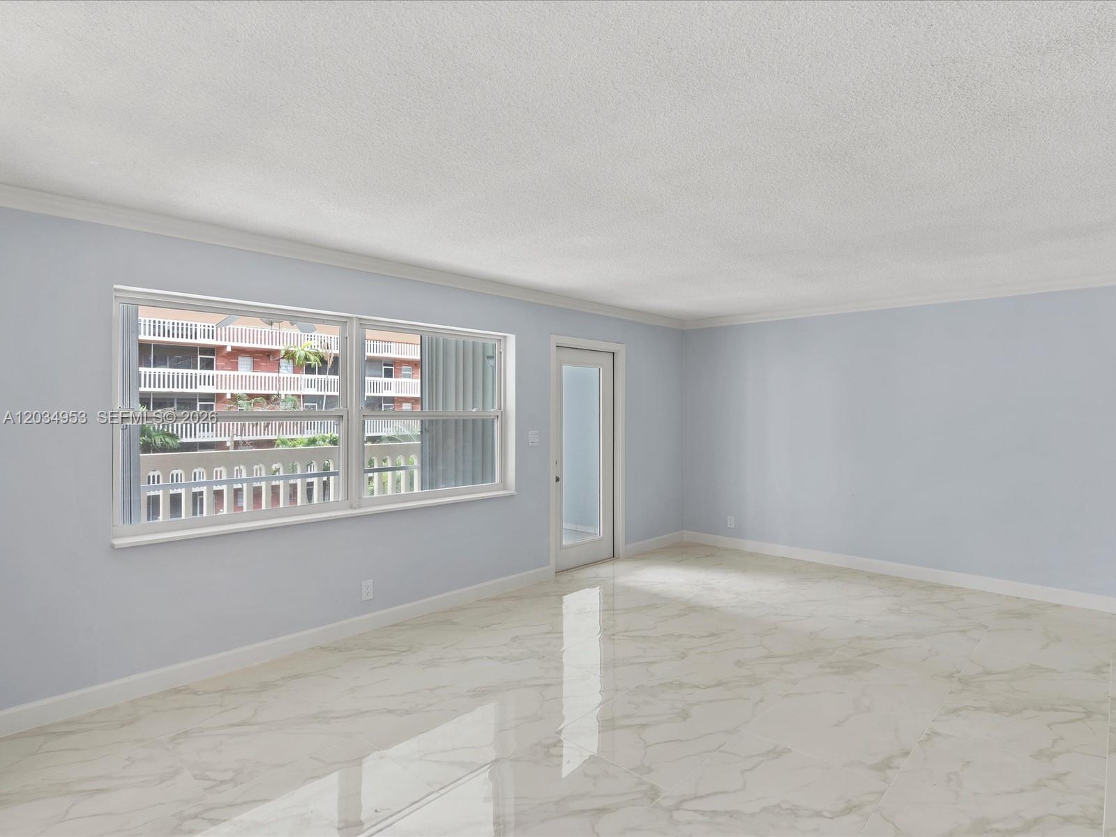 An empty room features light blue walls, white marble-look tile flooring, and a glass door — 200 NE 12th Ave # 7C, Hallandale Beach FL 33009, HALLANDALE BEACH, FL