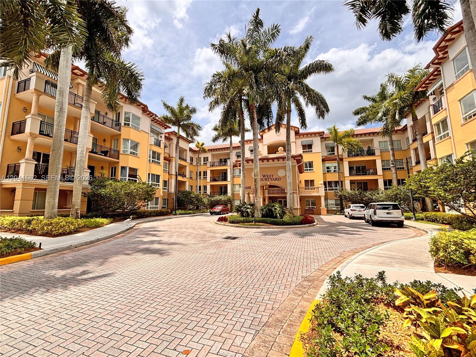 A paved circular driveway leads to the entrance of a multi-story yellow apartment building — 16101 Emerald Estates Dr # 155, Weston FL 33331, WESTON, FL