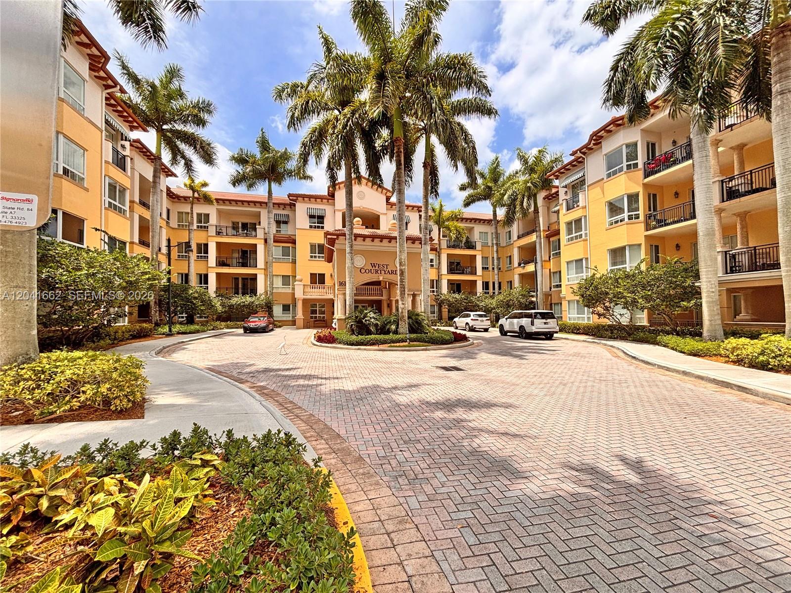 A paved circular driveway leads to the entrance of a multi-story yellow apartment building — 16101 Emerald Estates Dr # 155, Weston FL 33331, WESTON, FL