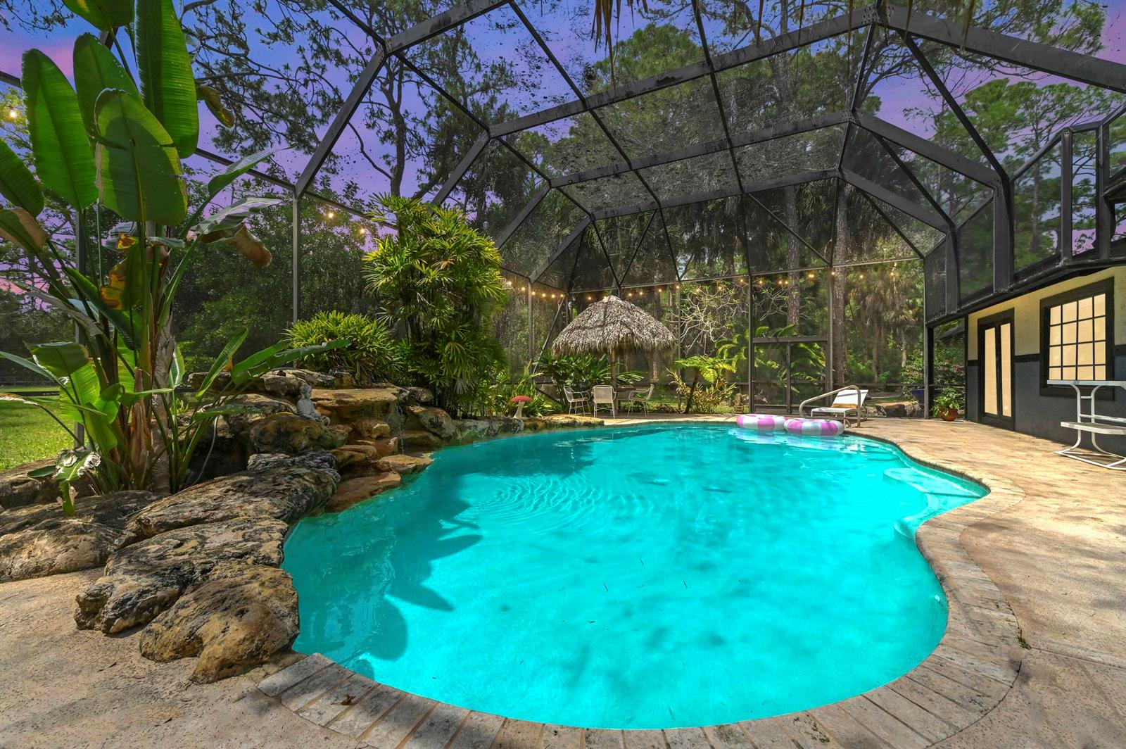 A screened-in swimming pool features a rock waterfall, tropical landscaping, and a thatched-roof tiki hut — 1339 Gallop Drive, Loxahatchee FL 33470, LOXAHATCHEE, FL